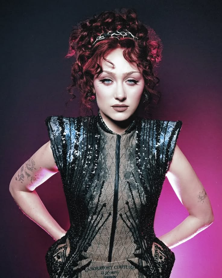 Person with curly hair and a headband stands with hands on hips wearing a sequined dress with structured shoulders and visible arm tattoos