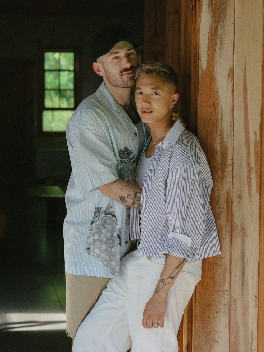 Two people standing close together indoors near a wooden wall with a window in the background