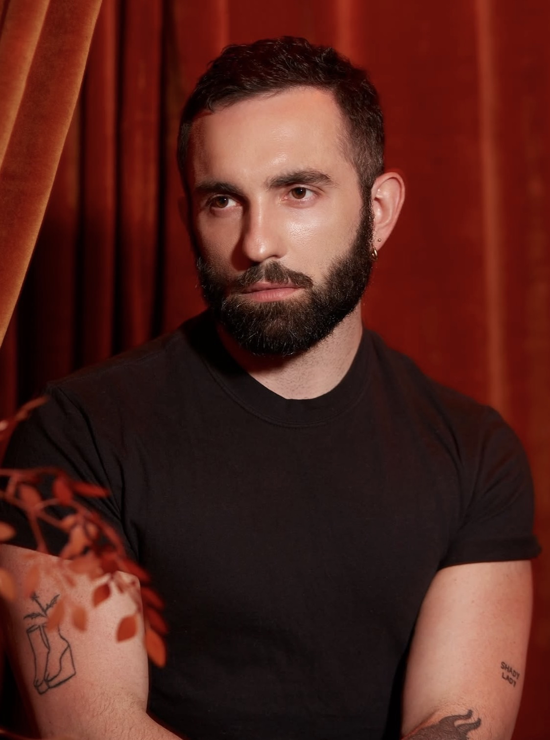 Peson with short hair waring a dark t-shirt and earrings, witting in front of a red curtain, with visible tattoos on both arms