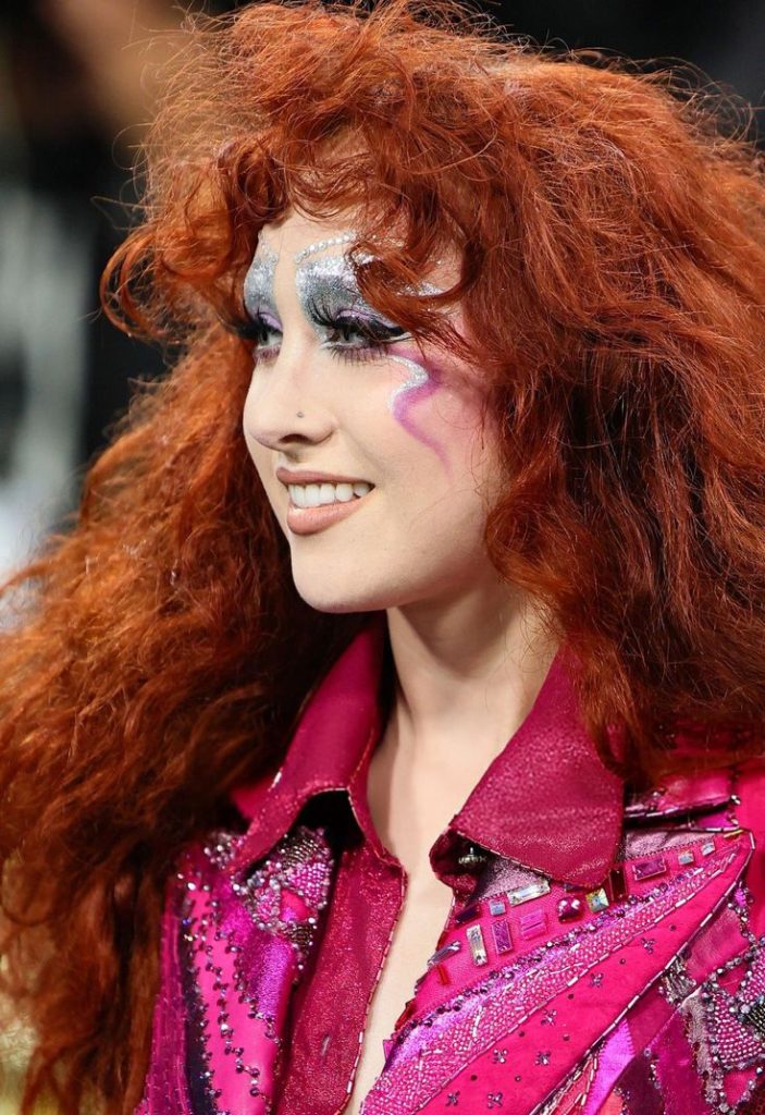 Person with voluminous curly hair wearing a pink studded jacket with a collar