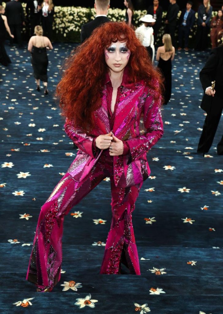 Person with long curly hair wearing a patterned suit standing on a dark carpet with floral designs