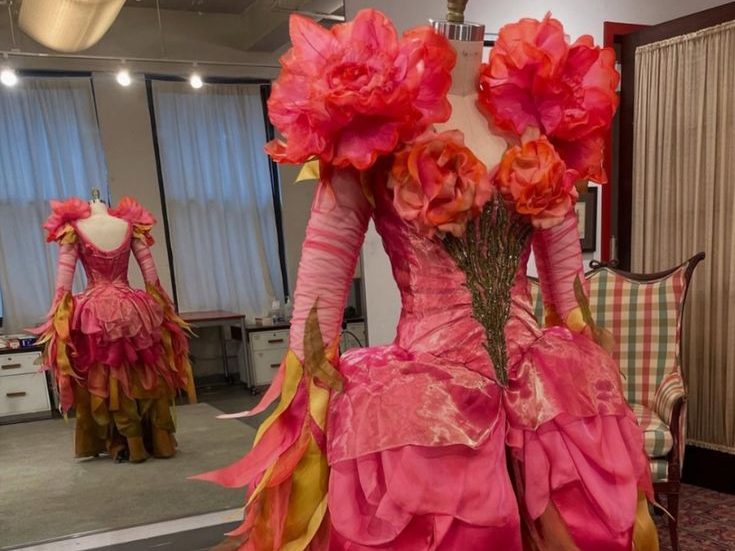 Mannequin wearing a pink and yellow gown with layered petal-shaped fabric and large rose-shaped decorations on the shoulders in front of a mirror in a room