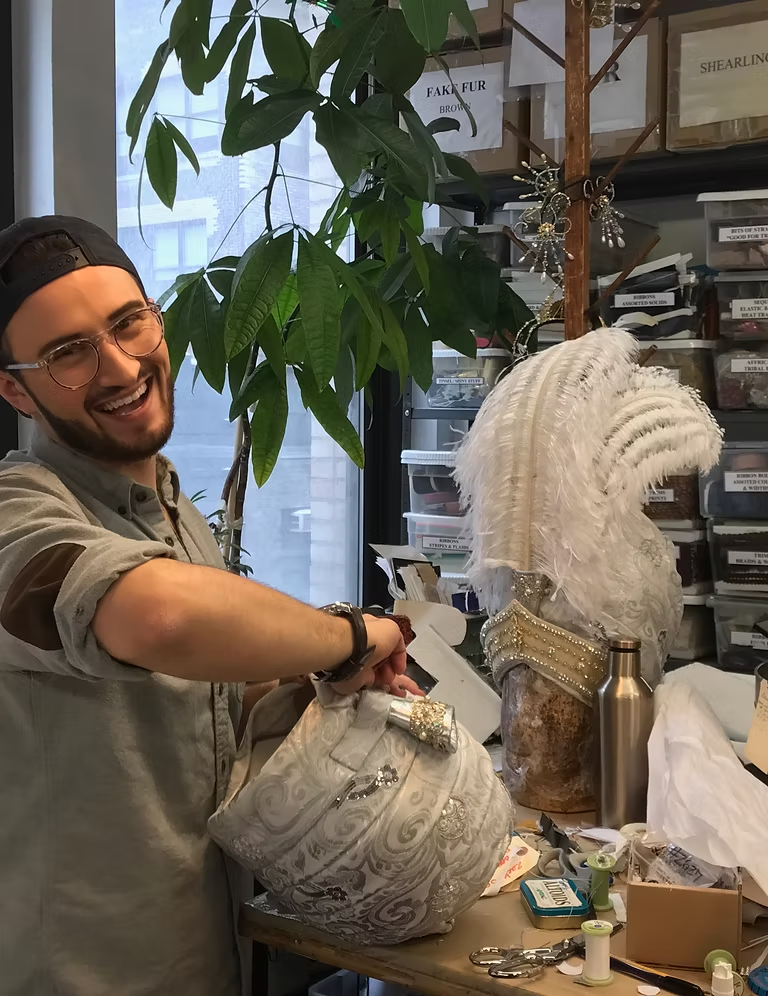 Person waring a light shirt and watch arranging white feathered costume pieces on a table in a cluttered room with a large green plant and a sign reading 'FAKE FUR' in the background