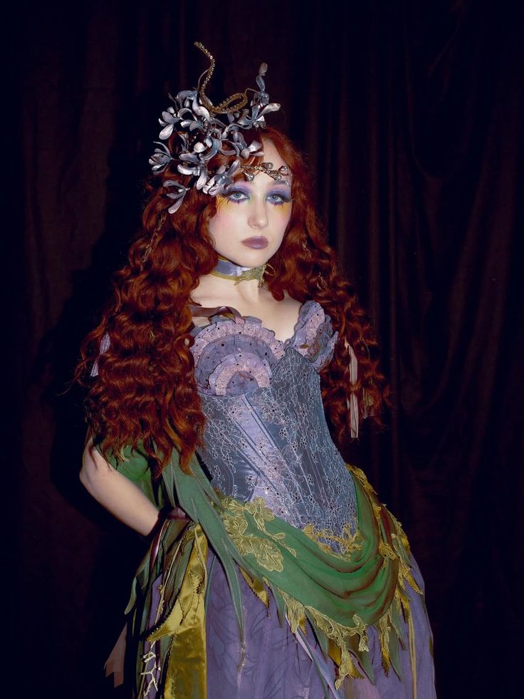 Person with long curly hair wearing a corset-style dress with green and pruple fabric and a headpiece made of twisted branches and flowers, posing against a dark background