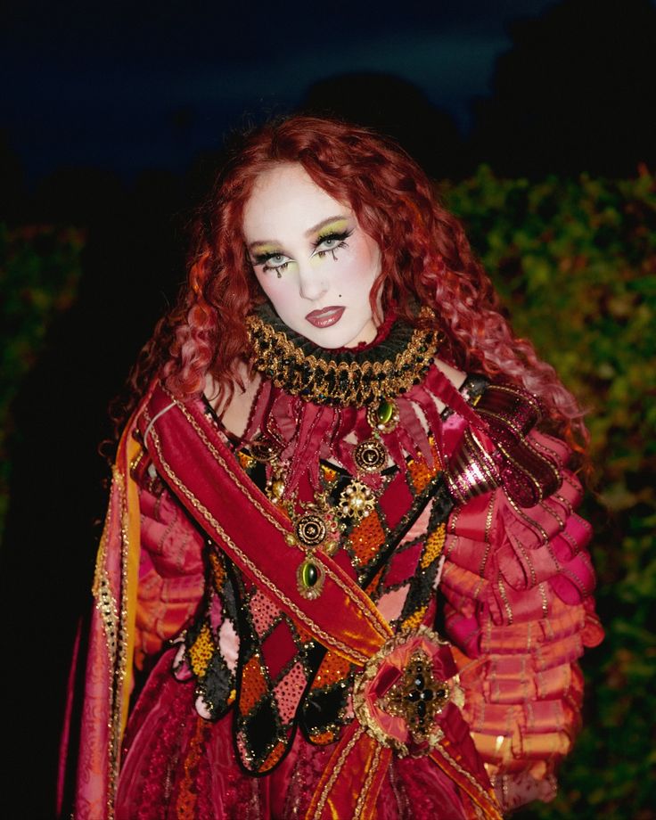 Person with long curly red hair wearing a red and black patterned costume with gold and green embellishments and dramatic makeup