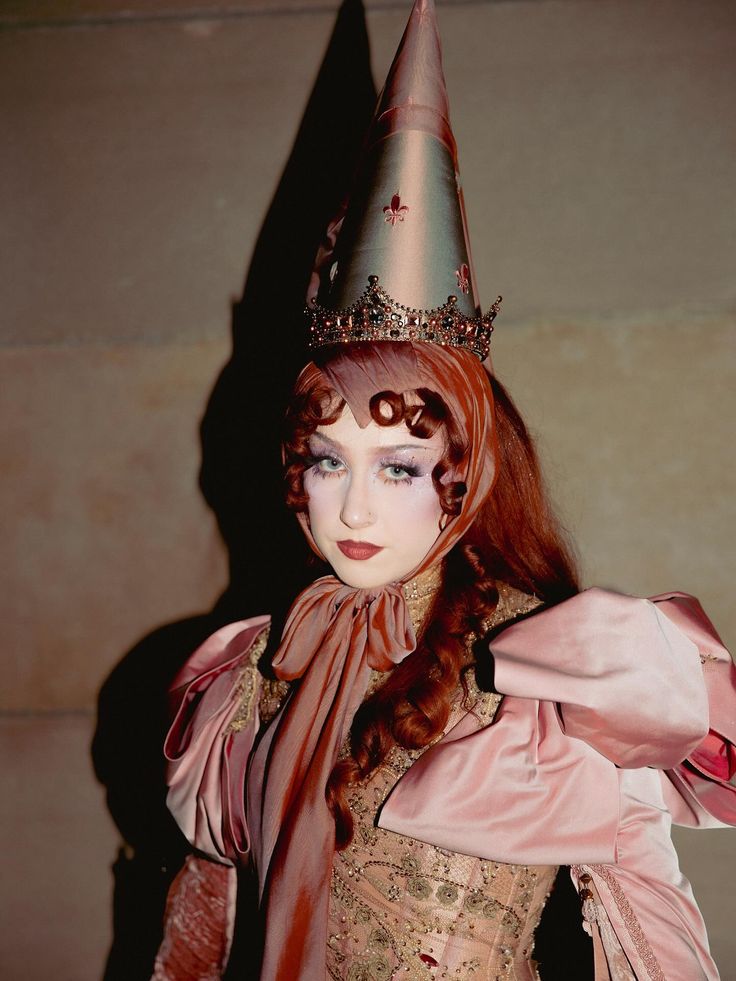 Person wearing a tall conical hat with a jeweled crown and a pink puffed-sleeve dress with a patterned bodice