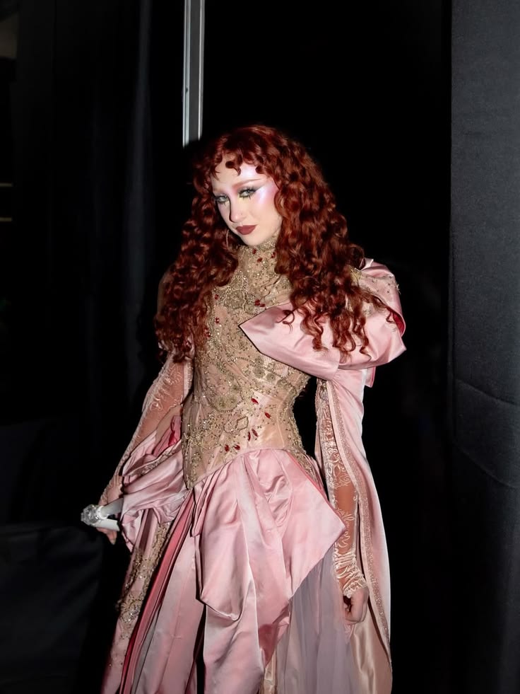 Person with long curly red hair wearing a detailed beige lace top and a voluminous pink skirt with draped fabric standing against a dark background