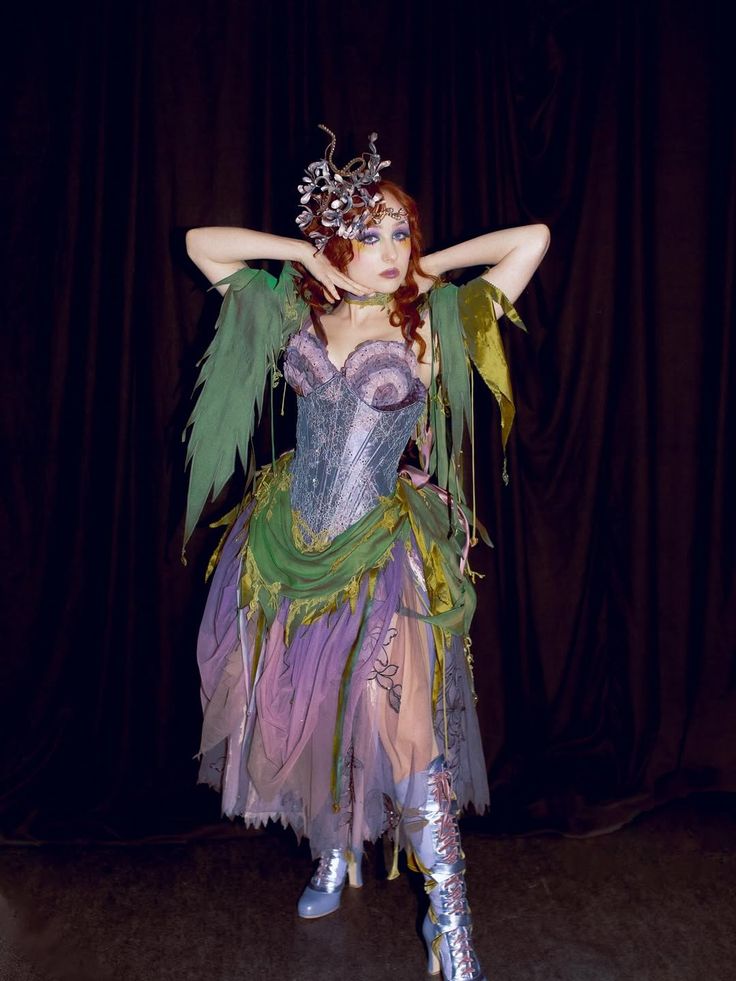 Person with long curly hair wearing a corset-style dress with green and pruple fabric and a headpiece made of twisted branches and flowers, posing with arms raised and hands near the face against a dark background