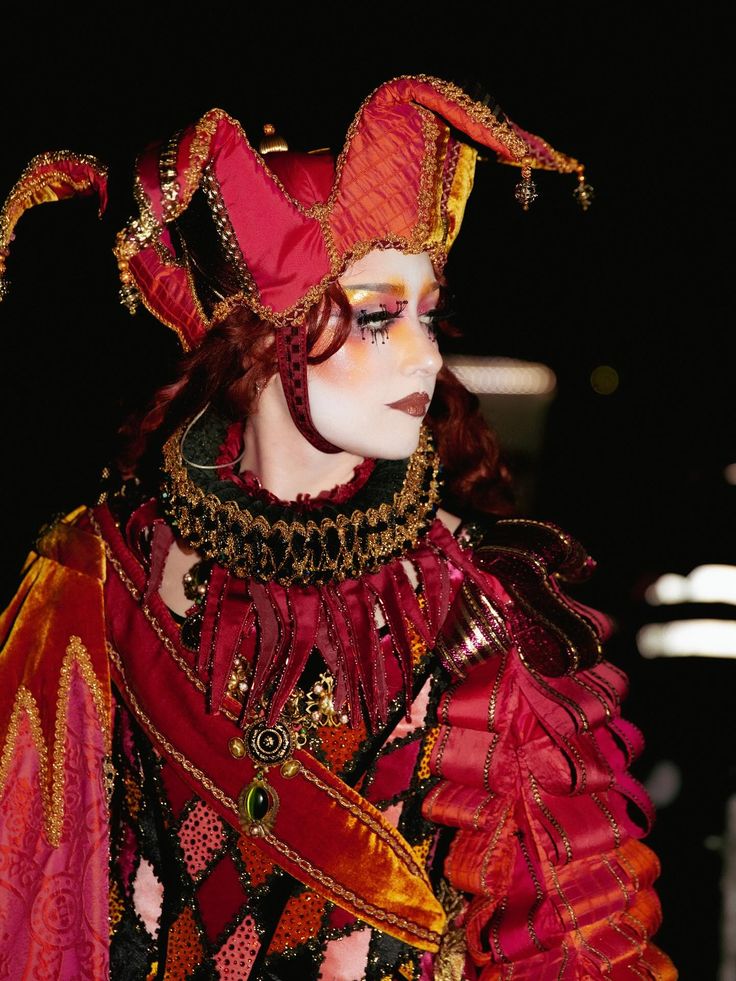 Person in elaborate jester costume with red and gold fabric, detailed collar, and face makeup looking to the side