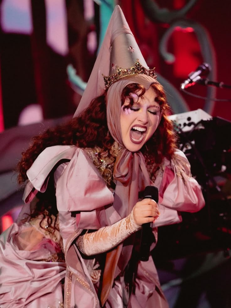 Chappell Roan singing passionately on stage wearing a pointed hat with a crown and a pink medieval-style dress with puffed sleeves
