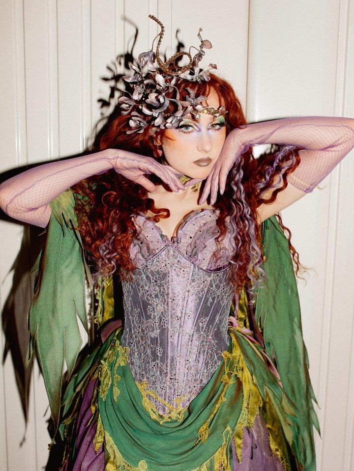 Person with long curly hair wearing a corset-style dress with green and pruple fabric and a headpiece made of twisted branches and flowers, posing with arms raised and hands near the face