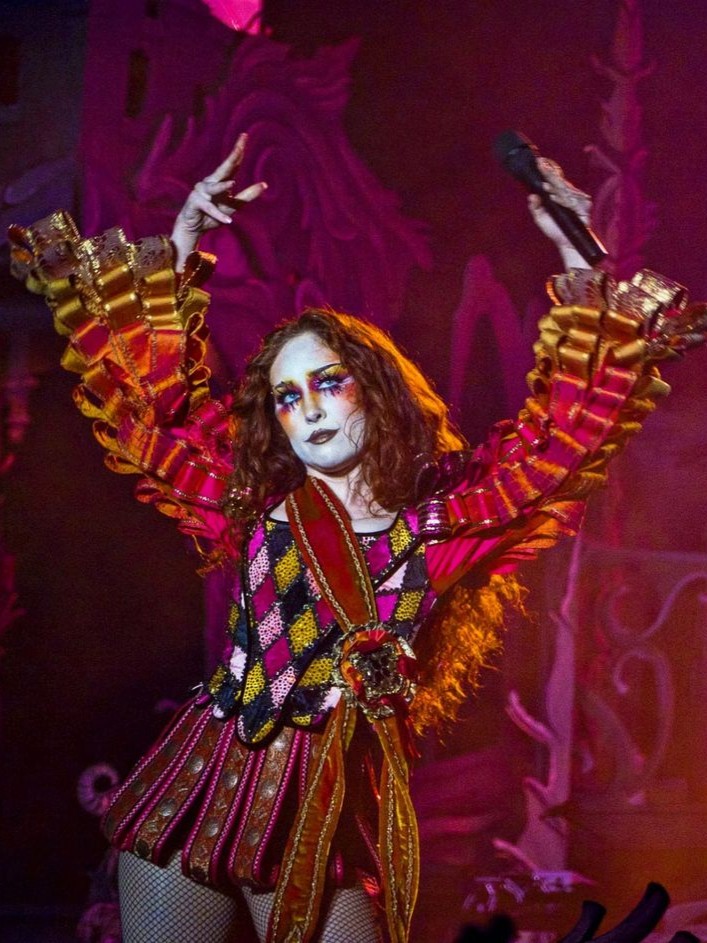 Person on stage wearing a colorful costume with ruffled sleeves and holding a microphone with arms raised