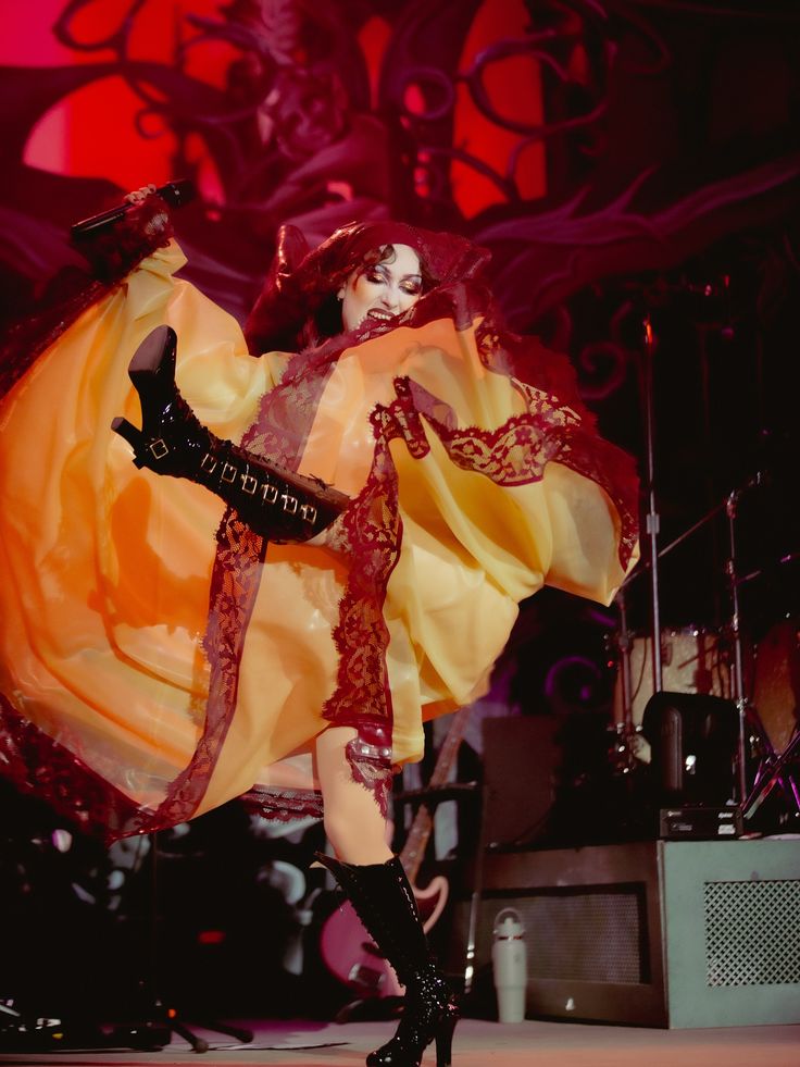 Person in a flowing, translucent gown with lace trim and high-heeled boots kicks one leg high on stage.