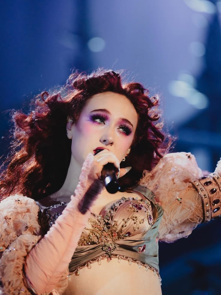 Person with curly hair wearing a detailed costume with puffed sleeves and holding a microphone on stage