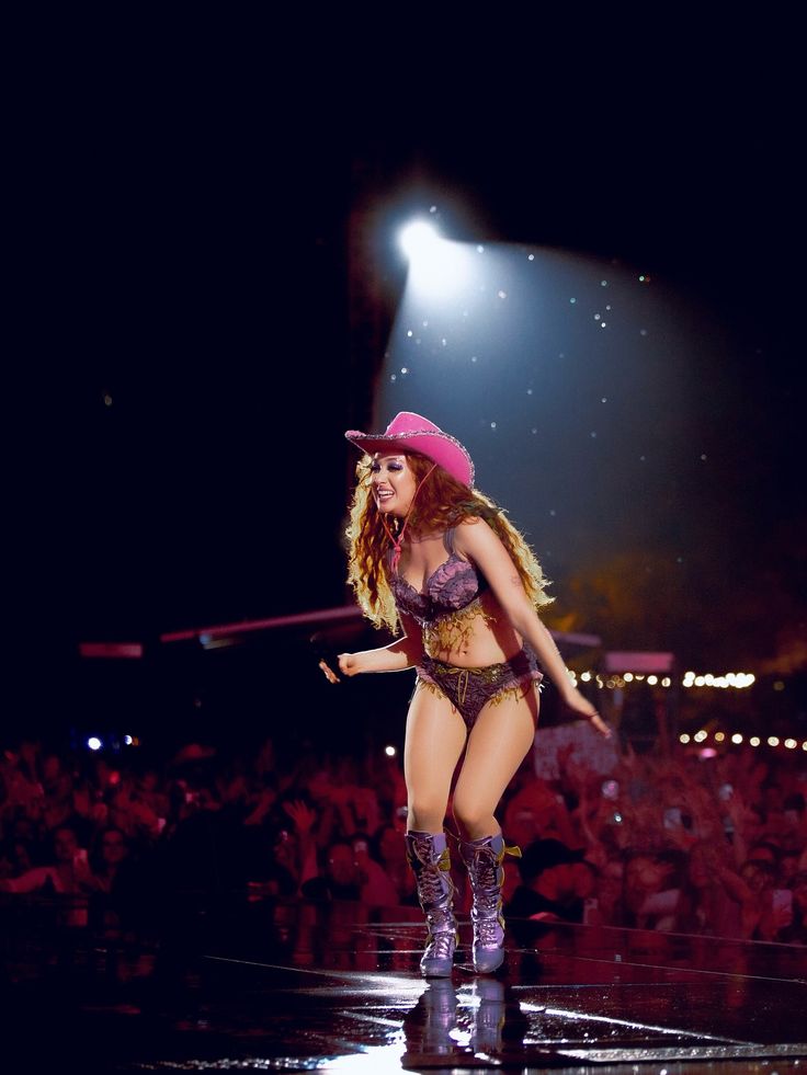 Woman with long curly hair wearing a purple bra and panties with a pink cowgirl hat on stage in front of a crowd