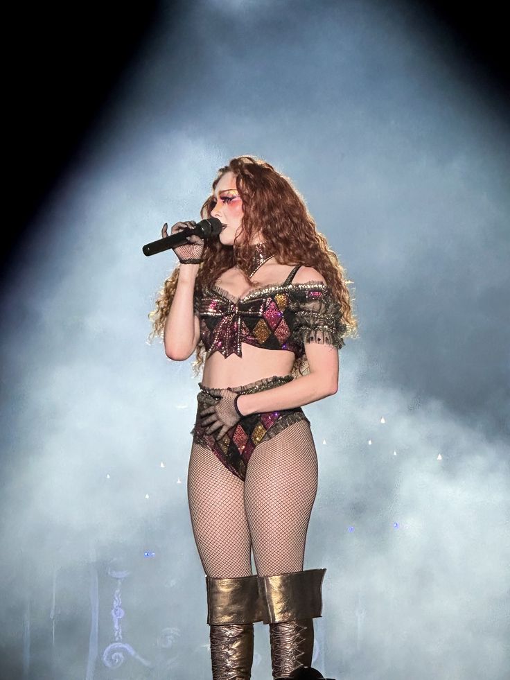 Woman with long curly hair wearing a patterned two-piece outfit and fishnet stockings singing into a microphone on stage with spotlight and smoke