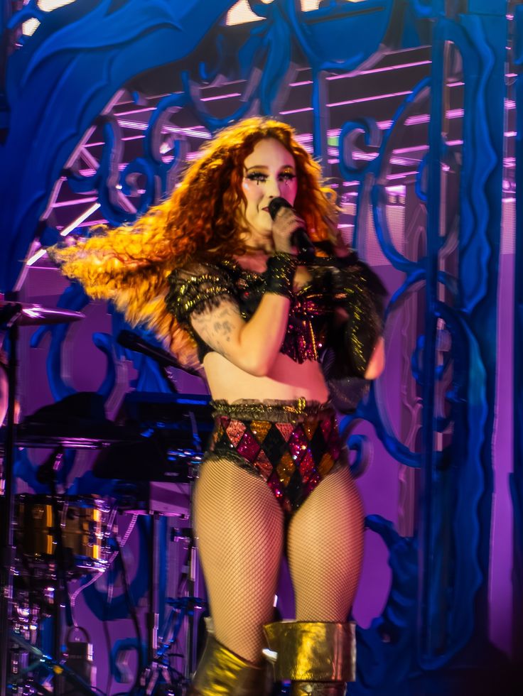 Performer with long curly hair wearing a sequined costume with fishnet stockings and gold boots on stage with decorative backdrop