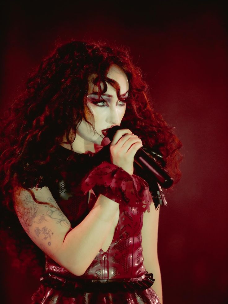 Person with long curly hair holding a microphone wearing a sleeveless red leather top and a red lace wristband