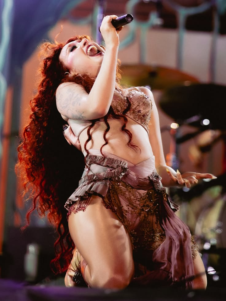 Female singer with long curly hair kneeling on stage holding a microphone wearing a sheer, embellished outfit