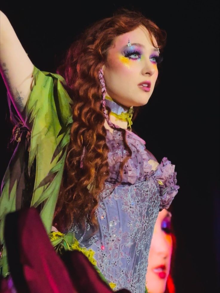 Person with long curly hair wearing a green and purple dress and vibrant make up standing on stage raising one arm