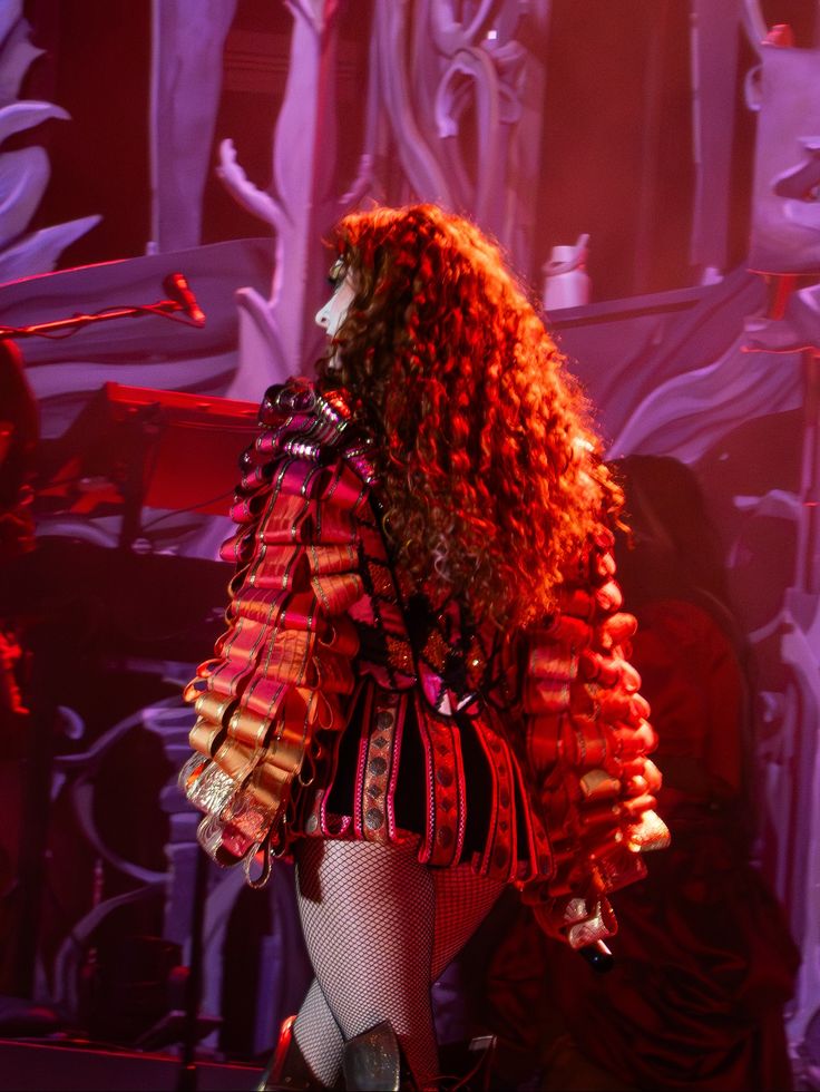 Person with long curly hair wearing a costume with large, layered, rectangular elements on the sleeves and fishnet stockings on stage