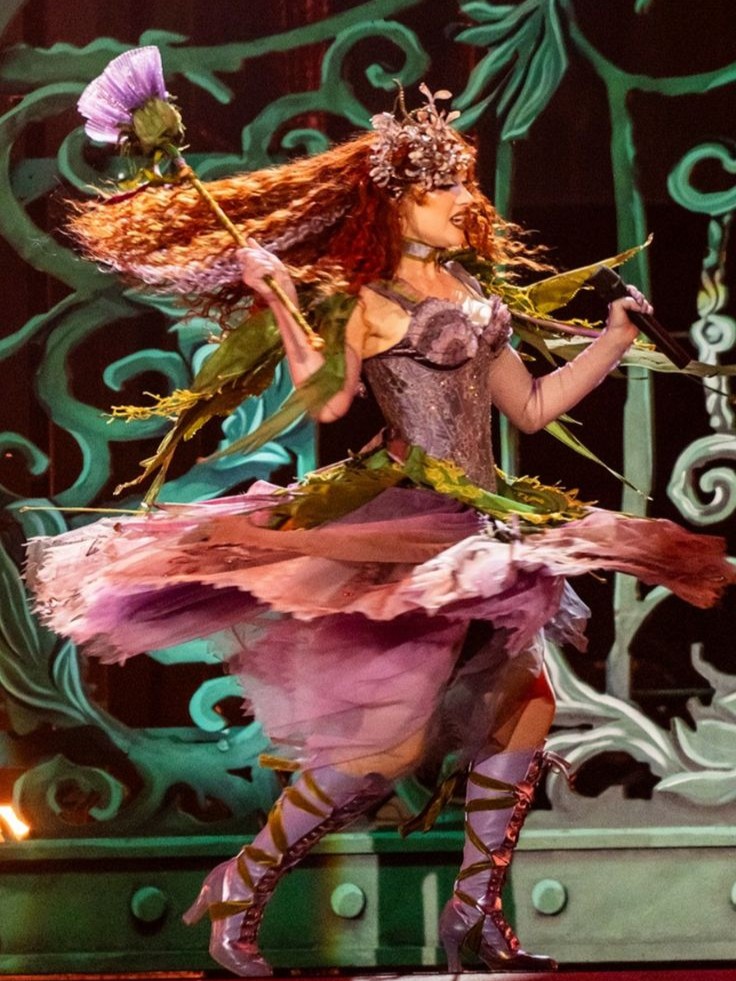 Woman in a fairy costume with a flower wand and a crown, wearing a layered skirt and lace-up boots, standing on stage against a decorative background
