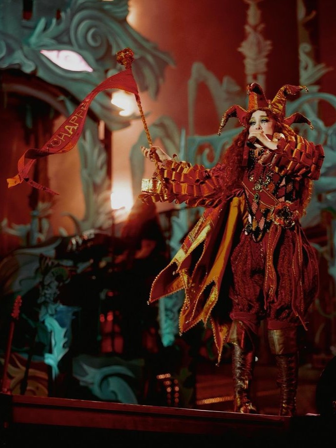 Figure dressed in a detailed jester costume holding a banner with text in a theatrical setting