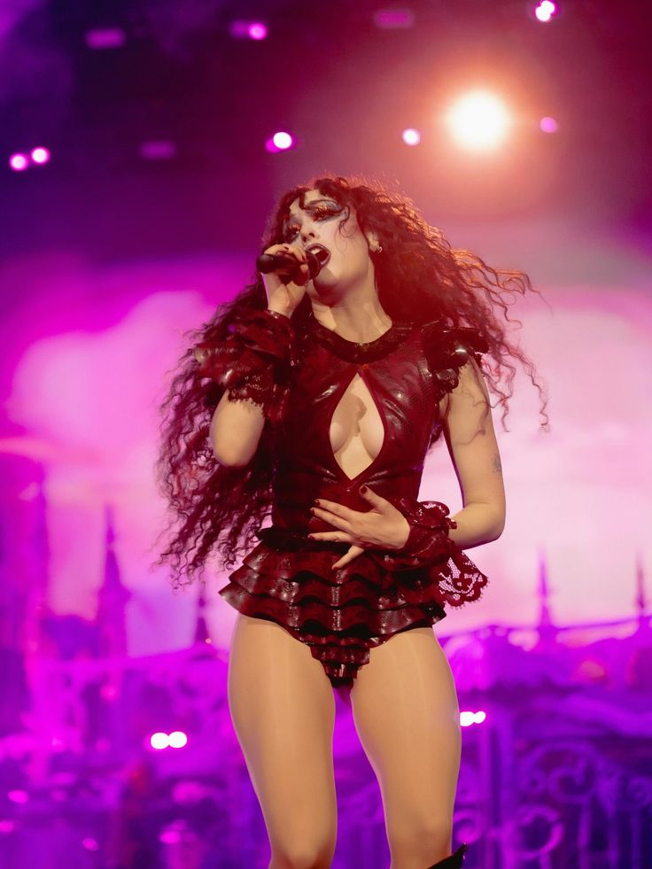 Female singer with long curly hair wearing a dark cutout outfit performing on stage with purple and pink lightning