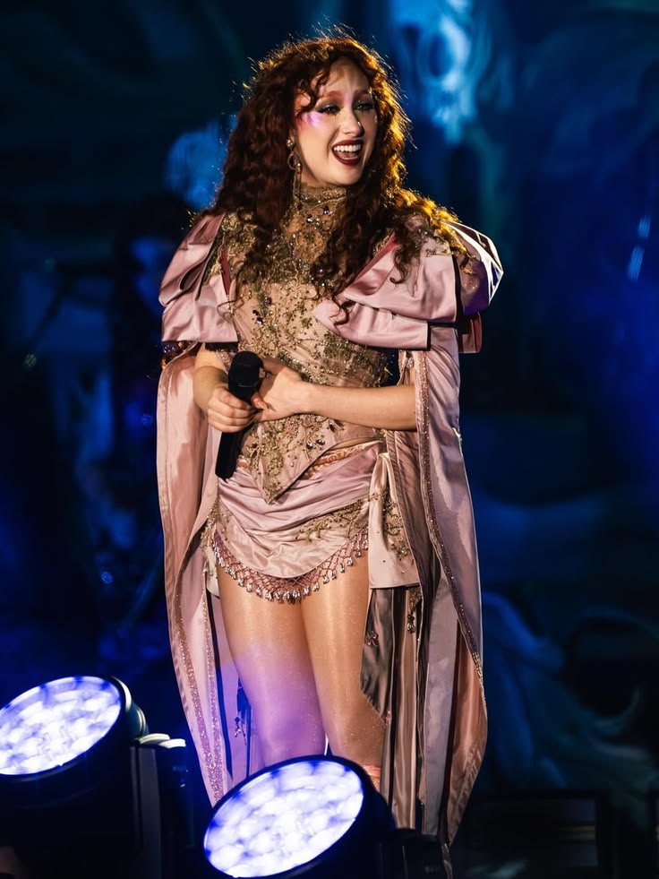 Woman with long curly hair wearing a detailed, layered stage costume holding a microphone under blue stage lights
