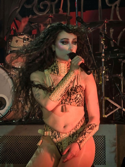 Femnale singer with long flowing hair wearing a patternes two piece outfit and holding a microphone on stage with a drum set in the background