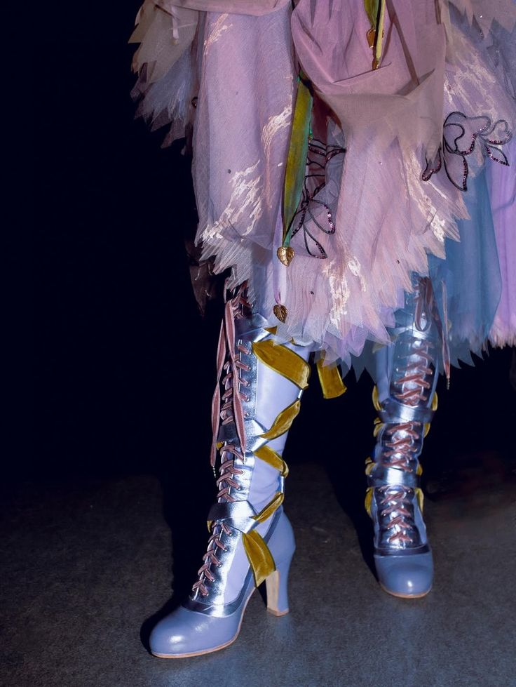 Pair of shiny lace up boots with gold ribbon details worn with a pleated skirt and a garment with floral embroidery
