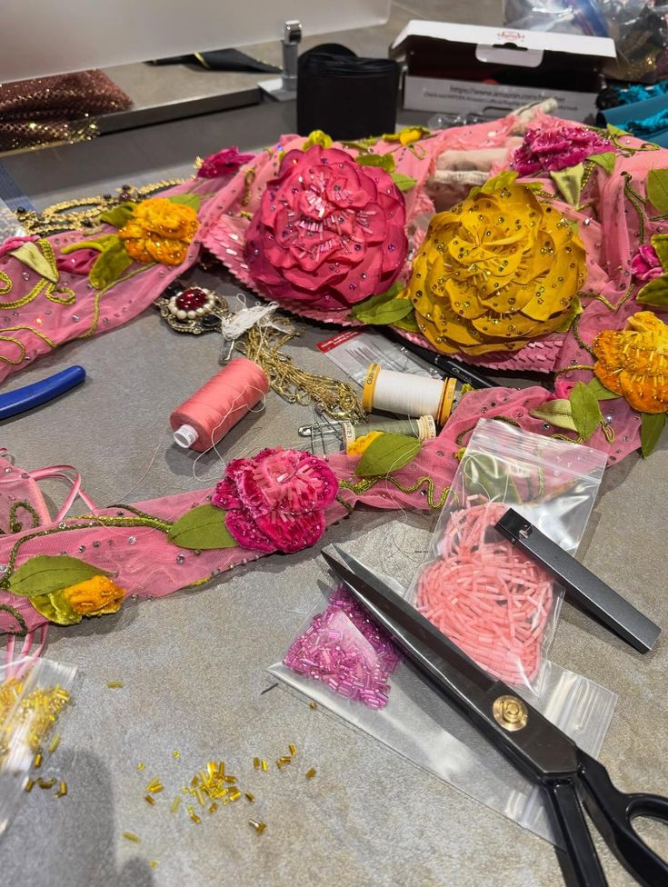 Table with pink fabric decorated with large yellow and pink fabric flowers, sewing threads, scissors, and small beads scattered around