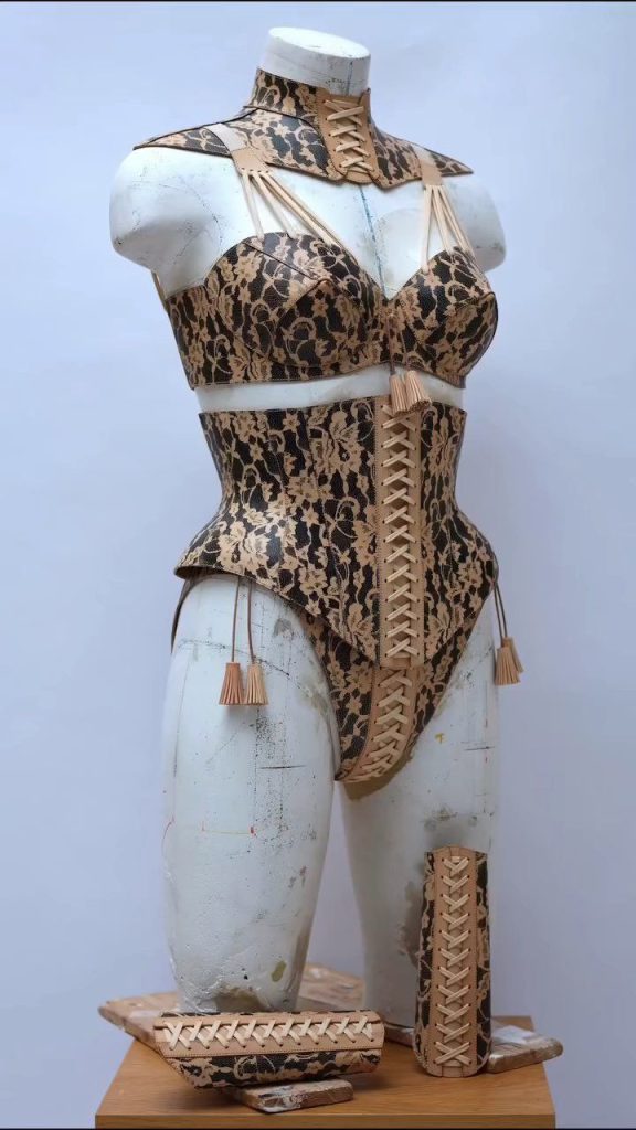 Mannequin wearing a lace-patterned set with a corset and matching high-waisted bottoms, displayed with a spool of thread and a lace piece on a wooden surface