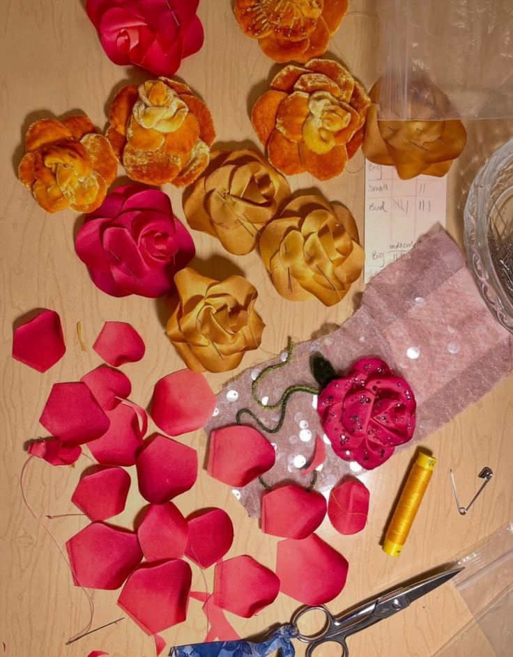 Multiple fabric flowers and loose fabric petals arranged on a table with a spool of yellow thread, scissors, a safety pin, and a piece of sequined fabric