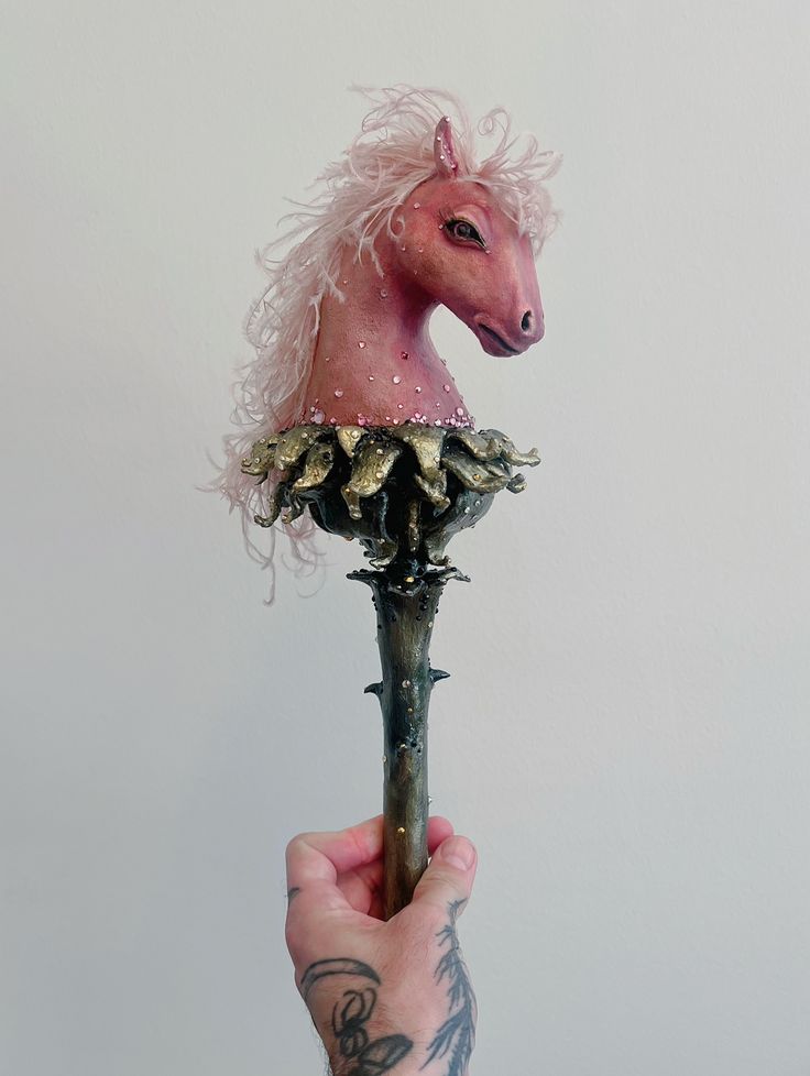 Hand holding a pink horse head sculpture with a curly mane mounted on a decorative metallic stand
