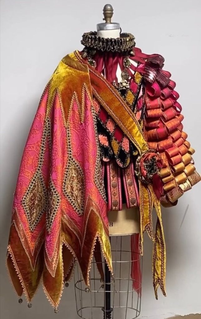 Mannequin dressed in a layered costume with a ruffled collar, a patterned cape with pointed edges, and a shoulder piece made of looped ribbons