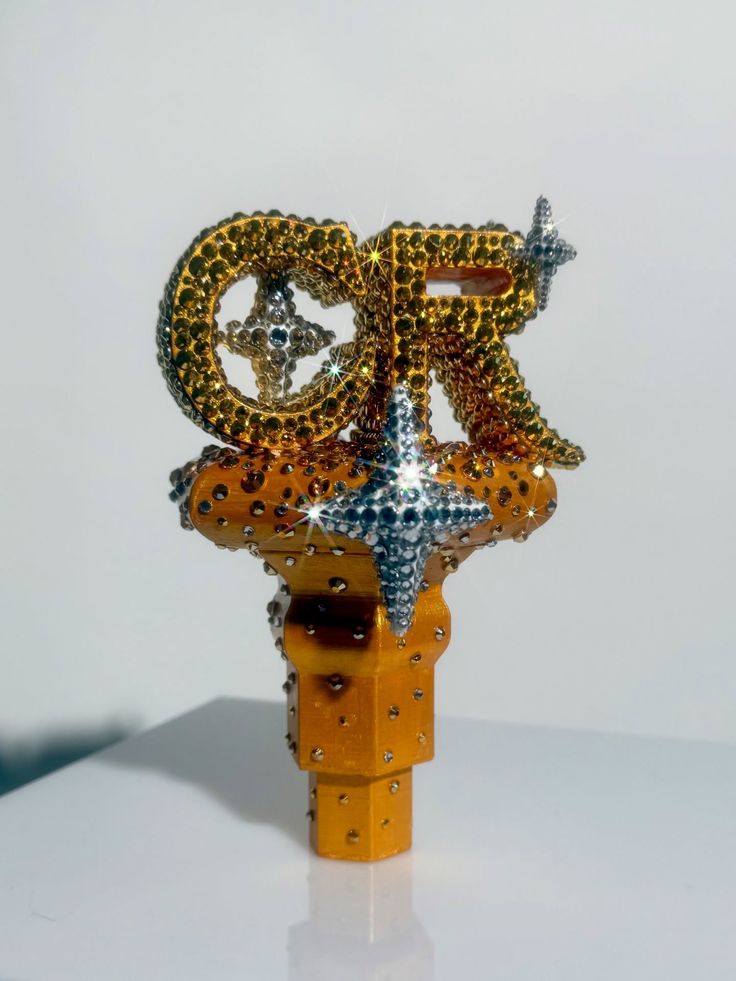 Decorative object shaped as the letters 'CR' adorned with gold and silver rhinestones on a yellow base