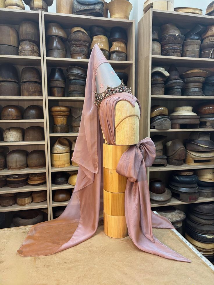 Medieval-style conical hat with long veil and jeweled band displayed on a wooden mannequin head in front of shelves filled with wooden hat blocks