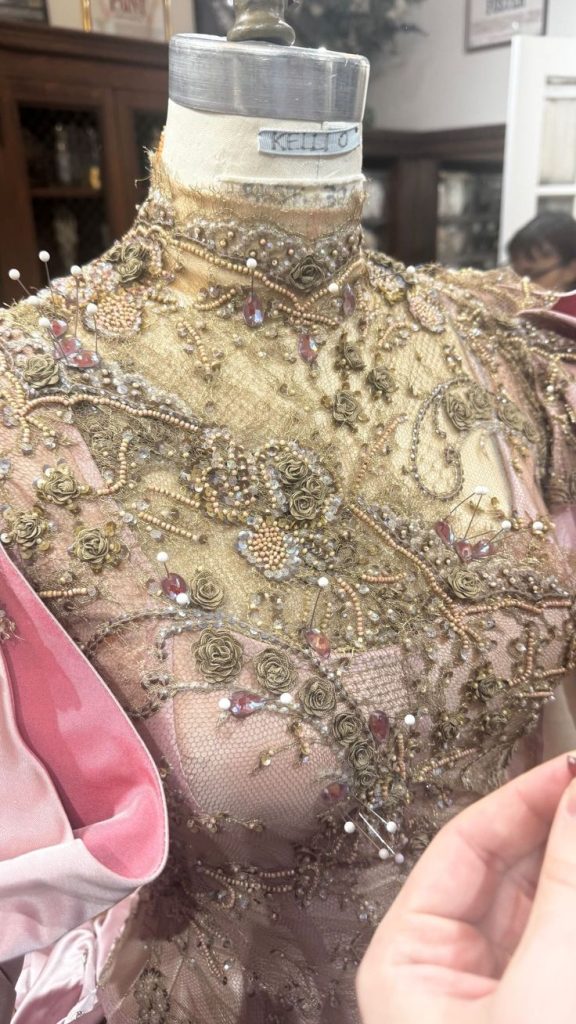 Close-up of a dressmaker's mannequin wearing a heavily beaded and embroidered garment with floral and geometric patterns and a pink fabric sleeve being adjusted by a hand